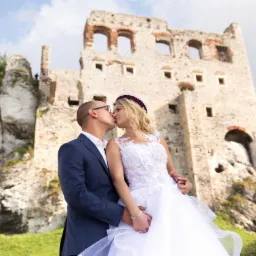 Pocałunek młodej pary na tle ruin zamku w Ogrodzieńcu, ona w koronkowej sukni ślubnej z tiulowym dołem, on w granatowym garniturze.