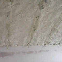 Biała, porowata izolacja natryskowa na poddaszu z widocznymi metalowymi łącznikami, ściana w tle w trakcie wykończenia.