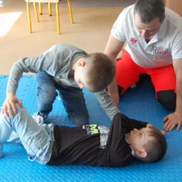 Instruktor w koszulce z logo udziela instrukcji dwóm chłopcom na niebieskiej macie edukacyjnej podczas zajęć z zakresu pierwszej pomocy.