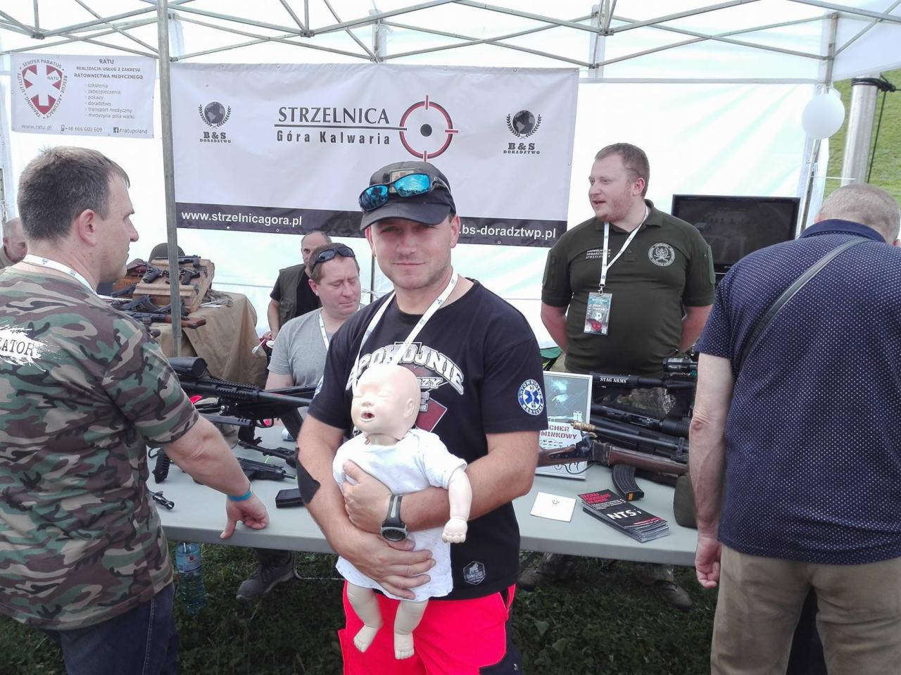 Instruktor z manekinem noworodka na tle stoiska strzelnicy z bronią, prawdopodobnie podczas demonstracji udzielania pierwszej pomocy w sytuacjach kryzysowych.