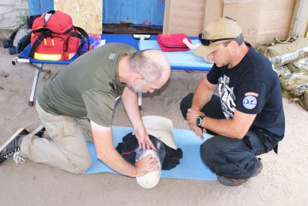 Instruktor demonstruje technikę uciskania klatki piersiowej na fantomie, drugi uczestnik obserwuje, w tle torba medyczna i apteczka.