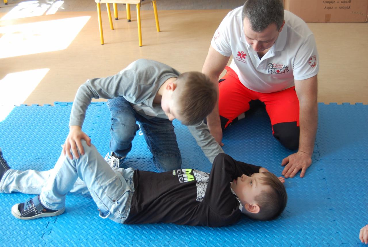 Instruktor w koszulce z logo udziela instrukcji dwóm chłopcom na niebieskiej macie edukacyjnej podczas zajęć z zakresu pierwszej pomocy.