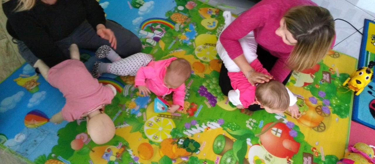 Instruktorka demonstruje technikę resuscytacji niemowlęcia na fantomie, obok leży niemowlę na kolorowej macie edukacyjnej.