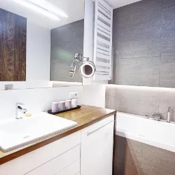 Nowoczesna łazienka z drewnianym blatem, szarą płytką imitującą kamień, białymi szafkami i podświetleniem LED pod wanną oraz dekoracyjnymi świecami.