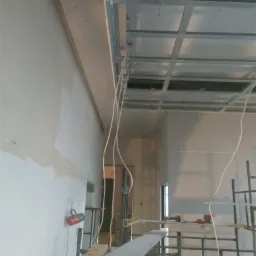 Wnętrze w trakcie prac wykończeniowych: widoczna konstrukcja sufitu podwieszanego z metalowych profili, częściowo obłożona płytami, instalacja elektryczna, fragment ściany po szpachlowaniu...