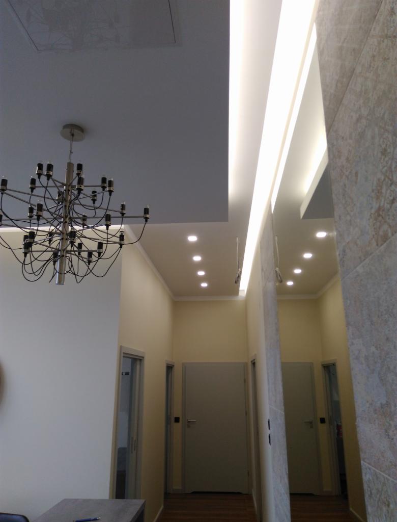 Korytarz z oświetleniem LED w suficie podwieszanym, ozdobnym żyrandolem i wykończeniem ścian w odcieniach beżu i szarości.