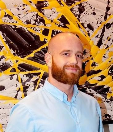 Portret mężczyzny z rudą brodą na tle abstrakcyjnego obrazu z czarnymi i żółtymi plamami.