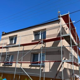 Usługi Ogólnobudowlane Bartosz Rospłoch - Dwukondygnacyjny dom z elewacją w odcieniach beżu, z widocznymi pasami w jaśniejszym i ciemniejszym kolorze, otoczony metalowymi rusztowaniami z czerwonymi pomostami, na tle błękitnego nieba.