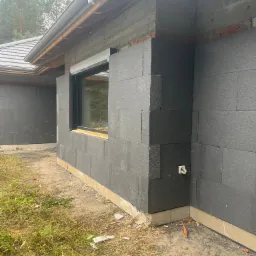 Dom w trakcie ocieplania: czarne płyty izolacyjne styropianowe na ścianach zewnętrznych, widoczne okno z zamontowaną roletą zewnętrzną, fragment dachu i rynny, teren wokół domu częściowo pokryty...