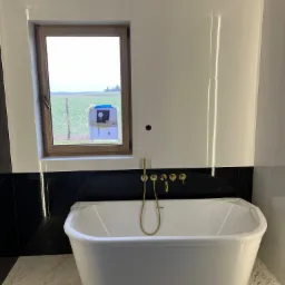 Nowoczesna łazienka w trakcie wykańczania: biała wanna wolnostojąca z złotą armaturą na tle białych płytek z czarnym pasem, widok na pole przez okno z zawieszonym rysunkiem domu.