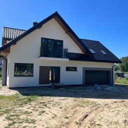 Usługi Ogólnobudowlane Bartosz Rospłoch - Nowoczesny dom jednorodzinny z balkonem i garażem, świeżo po malowaniu elewacji na jasny kolor, z ciemną dachówką i stolarką okienną. Widoczne puszki po farbie.