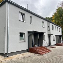 Usługi Ogólnobudowlane Bartosz Rospłoch - Szary budynek wielorodzinny po renowacji elewacji, z widocznymi rynnami i schodami wejściowymi. Jesienne drzewa w tle, pochmurne niebo.