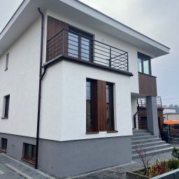 Usługi Ogólnobudowlane Bartosz Rospłoch - Nowoczesny dom z białą elewacją, drewnianymi akcentami i szarą podmurówką. Widoczny balkon z metalową balustradą i schody wejściowe. Minimalistyczny design.