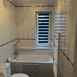 Usługi Ogólnobudowlane Bartosz Rospłoch - Jasna łazienka z wanną, toaletą i grzejnikiem drabinkowym. Widoczne płytki z dekoracyjnym pasem i okno z roletami dzień-noc. Częściowo widoczne narzędzia.