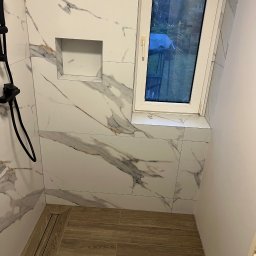 Usługi Ogólnobudowlane Bartosz Rospłoch - Wykończona kabina prysznicowa z marmurowymi płytkami, wnęką na kosmetyki i odpływem liniowym. Widoczne okno dachowe i drewnopodobna podłoga.