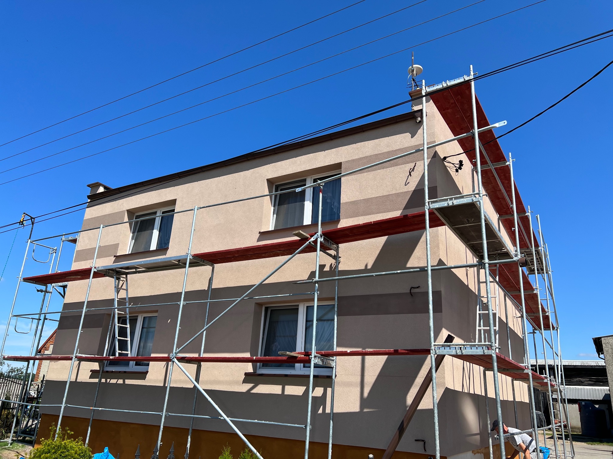 Dwukondygnacyjny dom z elewacją w odcieniach beżu, z widocznymi pasami w jaśniejszym i ciemniejszym kolorze, otoczony metalowymi rusztowaniami z czerwonymi pomostami, na tle błękitnego nieba.