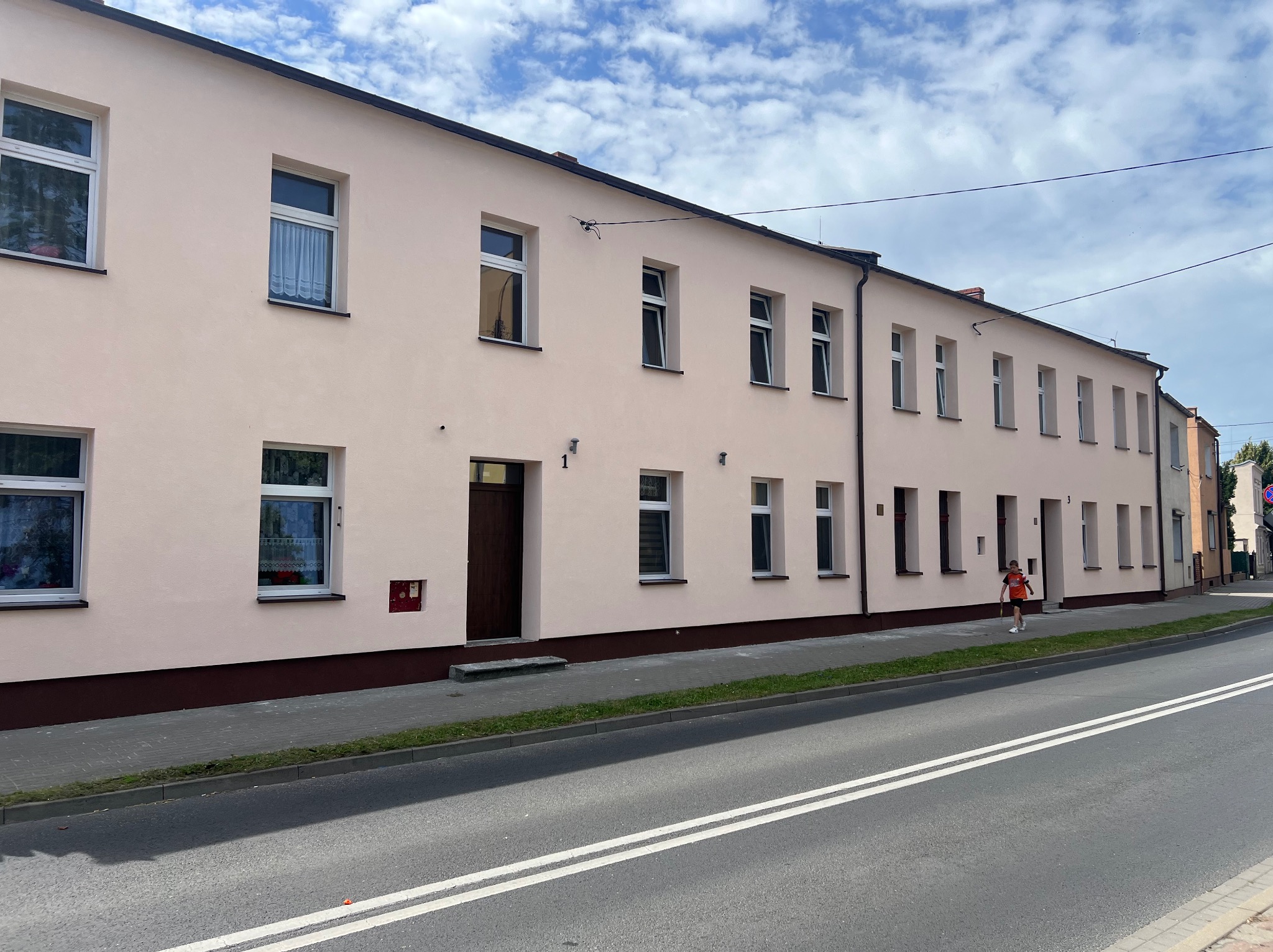 Długi budynek mieszkalny z jasnoróżową elewacją i białymi oknami, widoczny z poziomu ulicy w słoneczny dzień z lekkim zachmurzeniem.