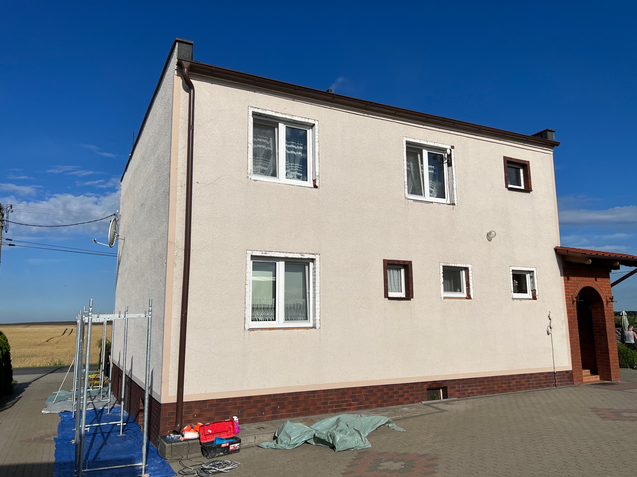Dwukondygnacyjny dom z beżową elewacją, widoczne rusztowanie, narzędzia i plandeka, wskazujące na prace elewacyjne w toku, z polem uprawnym w tle.