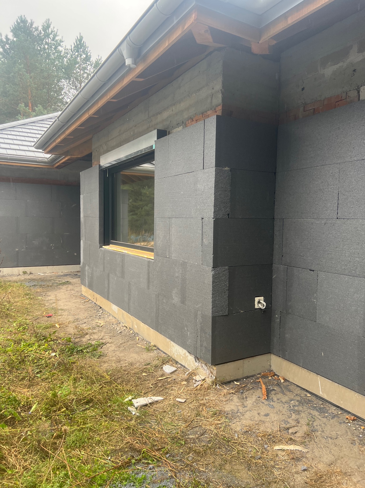 Dom w trakcie ocieplania: czarne płyty izolacyjne styropianowe na ścianach zewnętrznych, widoczne okno z zamontowaną roletą zewnętrzną, fragment dachu i rynny, teren wokół domu częściowo pokryty...