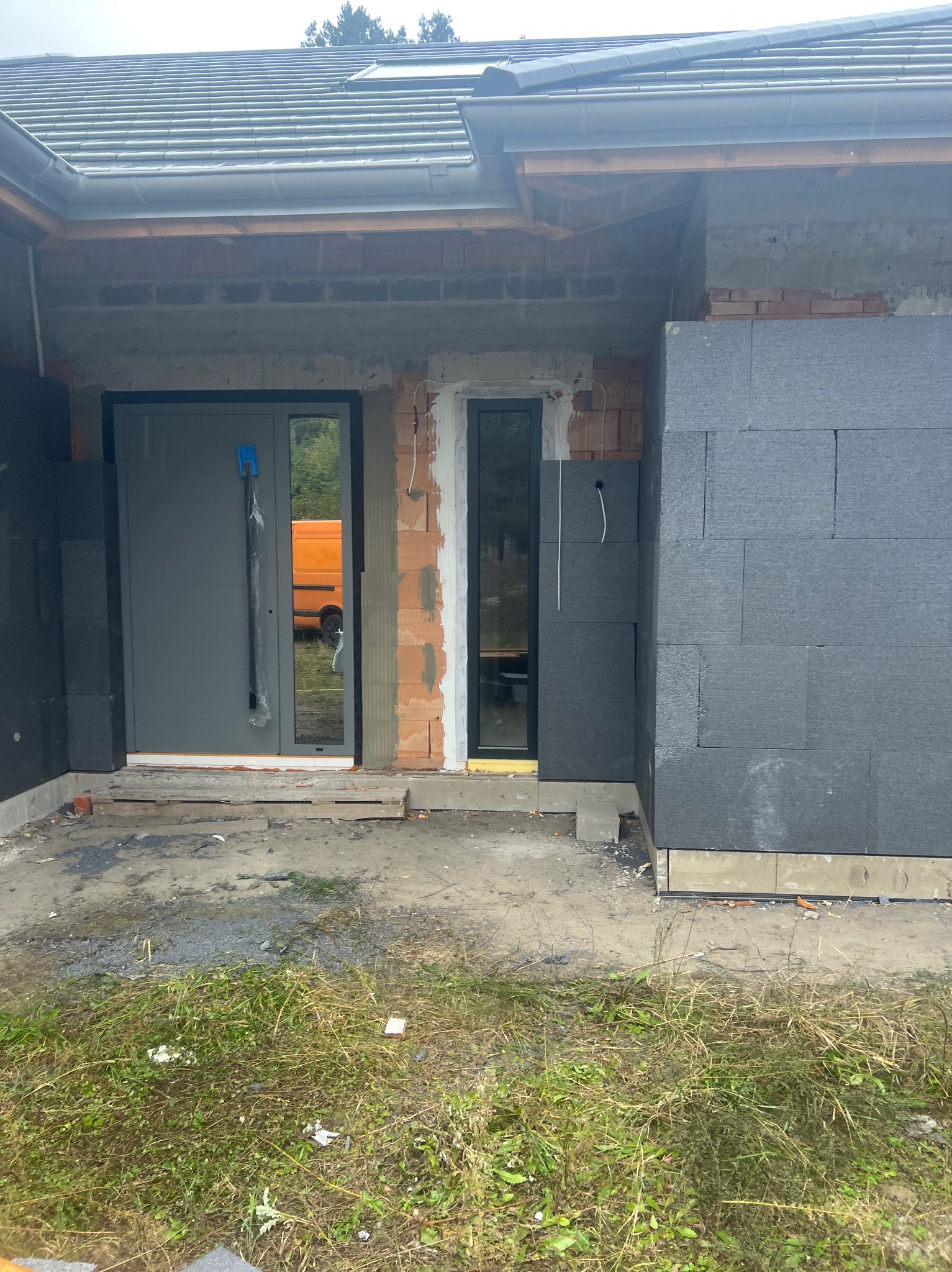 Ocieplanie elewacji domu jednorodzinnego z szarym styropianem, widoczna nowa stolarka okienna i drzwiowa, teren wokół w trakcie prac wykończeniowych.