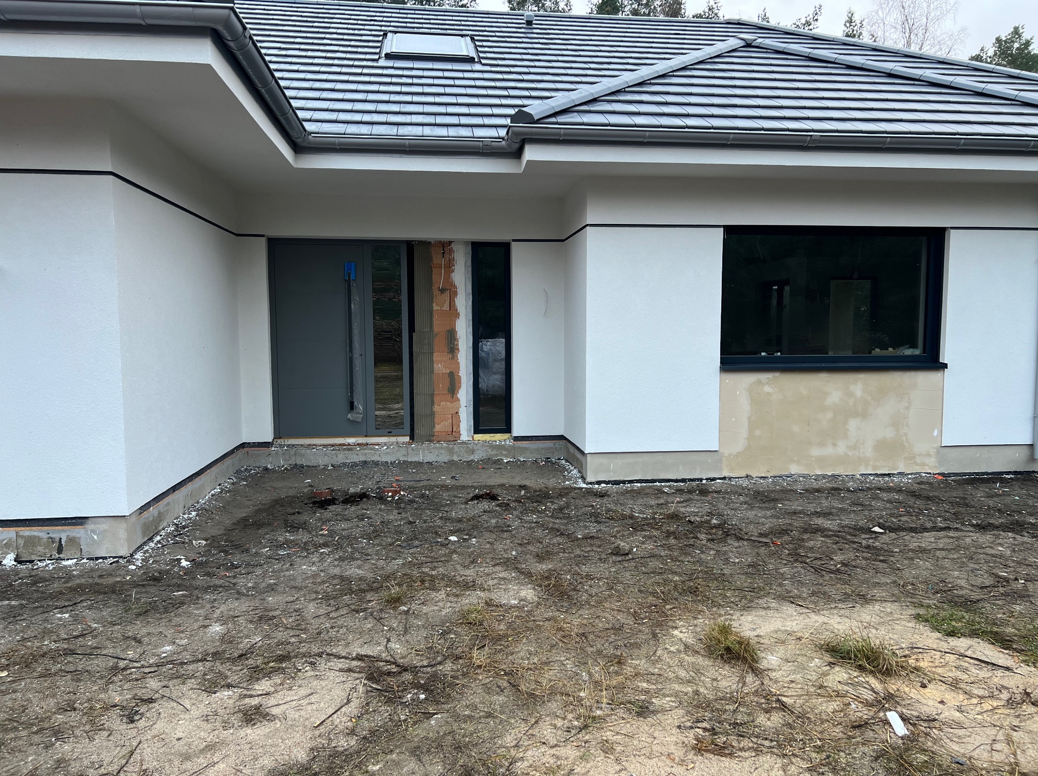 Nowoczesny dom jednorodzinny w trakcie wykończenia elewacji, widoczna surowa ściana obok okna, niezabezpieczone wejście i niezagospodarowany teren wokół budynku.