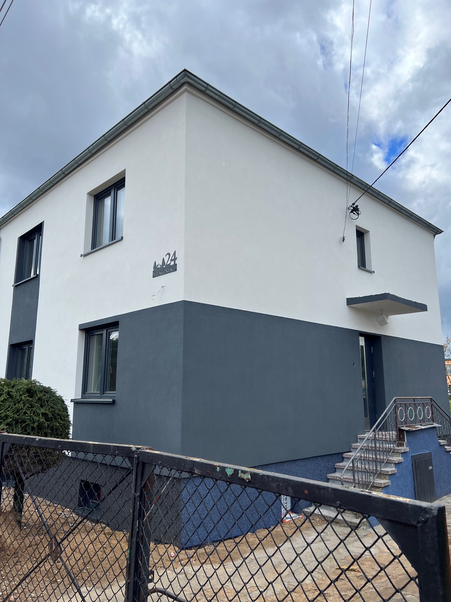 Dwukondygnacyjny dom z elewacją w kolorach szarości i bieli, numer posesji '24' z motywem drzew iglastych, widok zza metalowego ogrodzenia.
