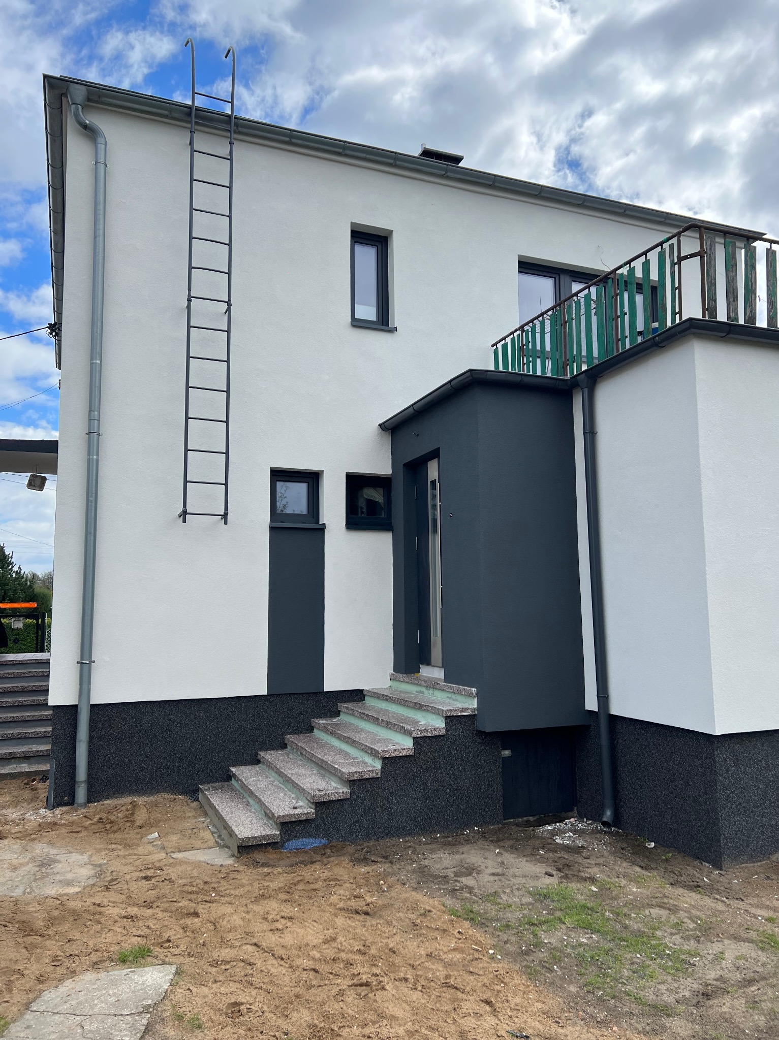 Dwukondygnacyjny dom z białą elewacją i ciemnoszarymi akcentami, schody wejściowe wyłożone kamieniem, metalowa drabina przymocowana do ściany, balkon z zieloną balustradą, teren wokół domu...