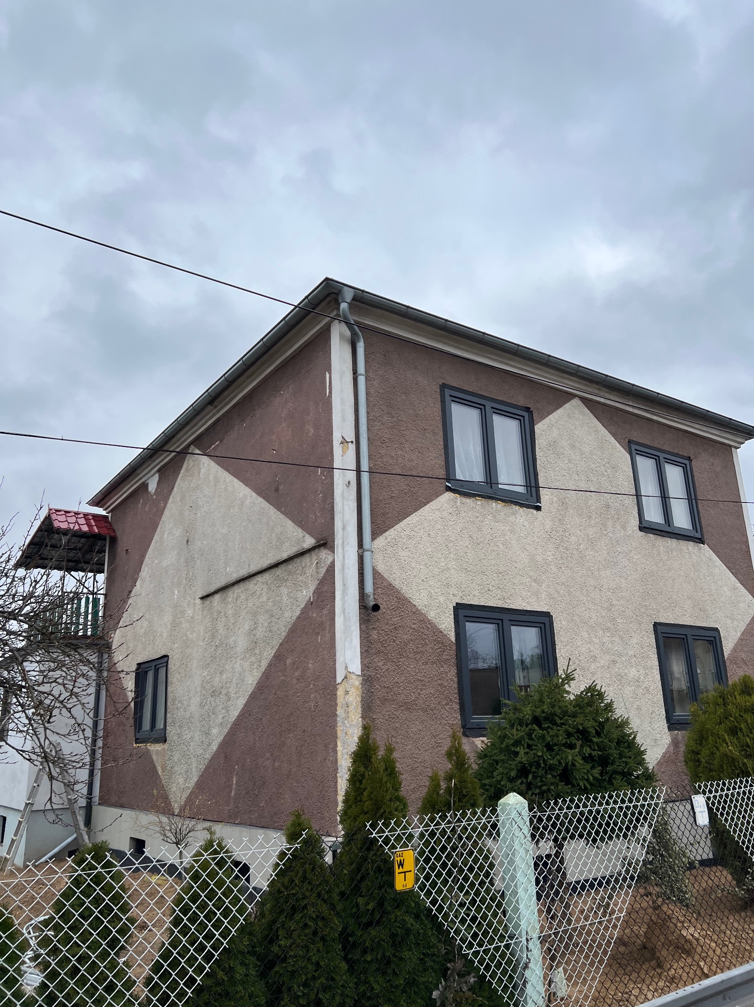 Dwukondygnacyjny dom z elewacją w geometryczne wzory (brązowo-beżowe romby), ciemne ramy okienne, widok z dołu, ogrodzenie z siatki na pierwszym planie.