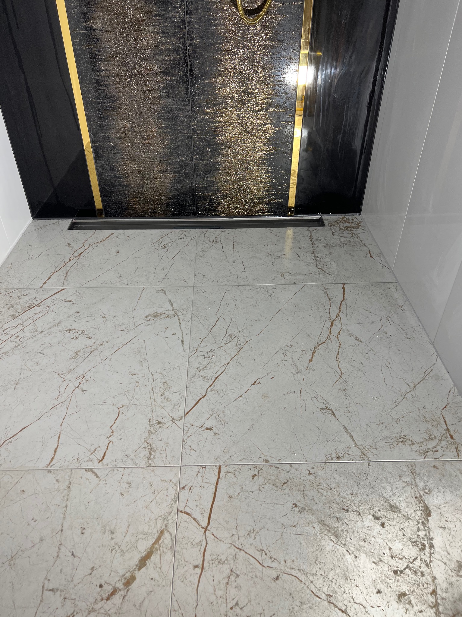 Nowoczesna kabina prysznicowa z czarną ścianą akcentową z brokatem, złotymi paskami i odpływem liniowym w podłodze z płytek imitujących marmur.