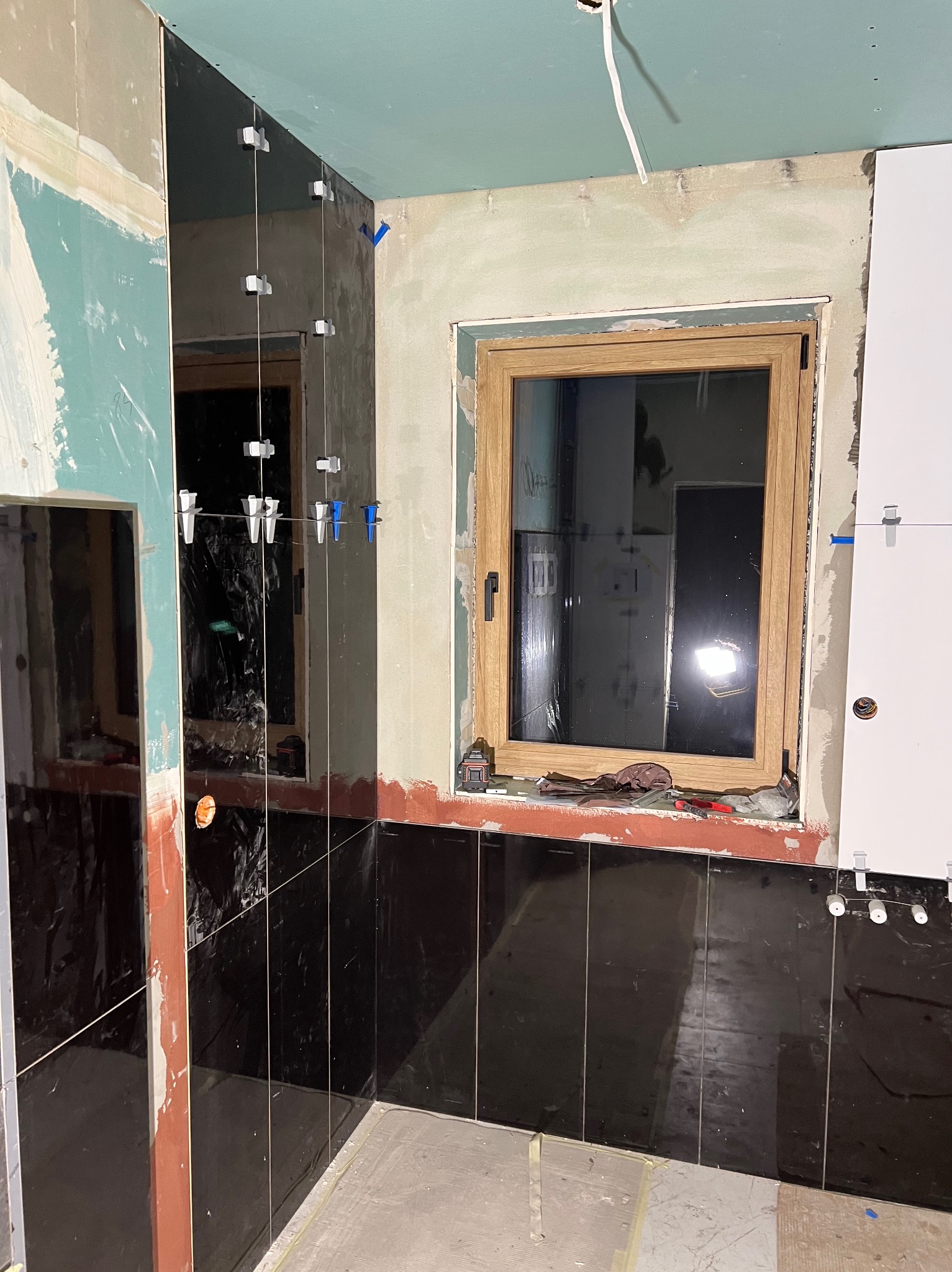 Wnętrze łazienki w trakcie układania czarnych, błyszczących płytek ściennych, widoczne plastikowe krzyżyki dystansowe i narzędzia na parapecie okna.