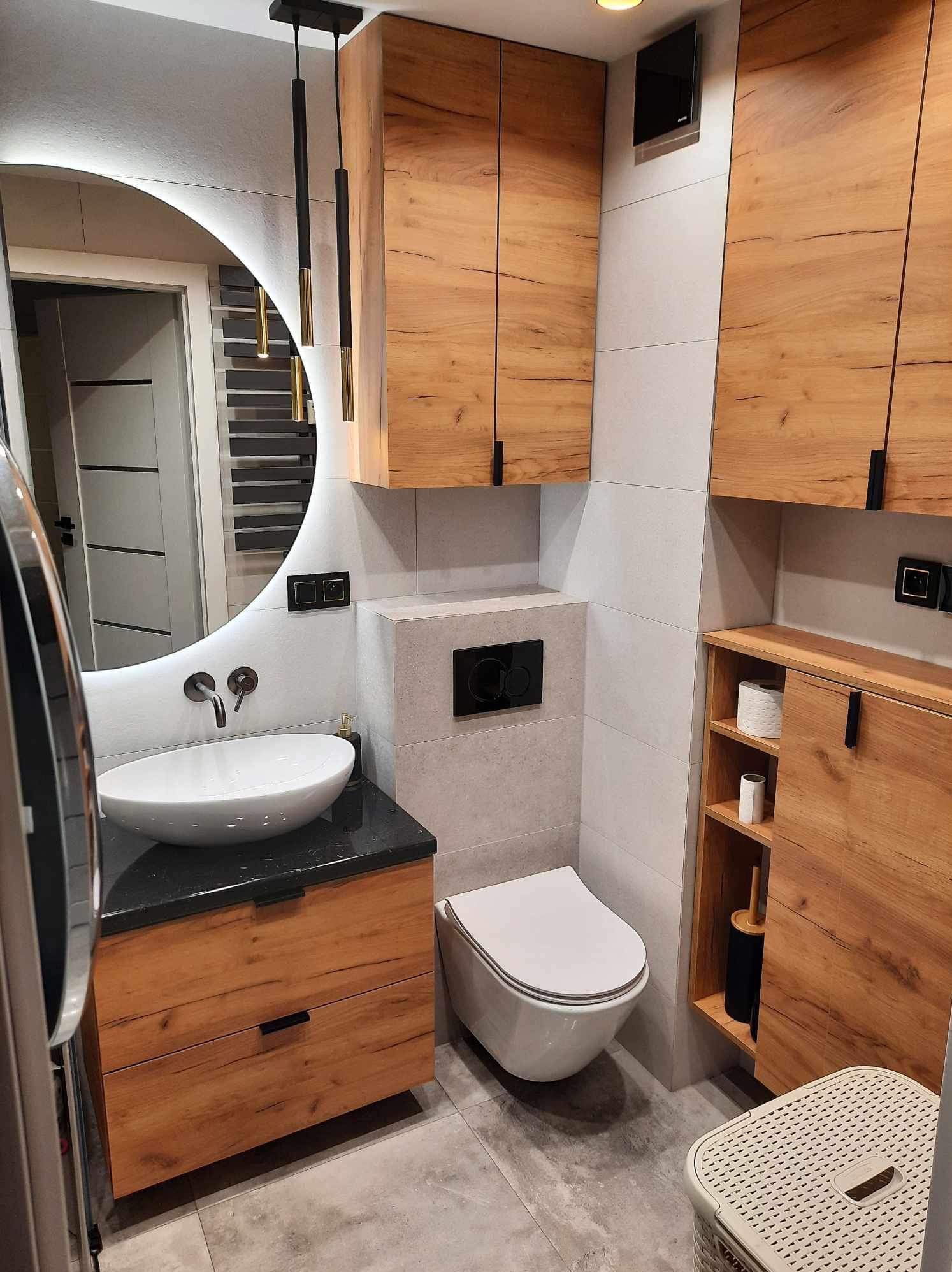 Nowoczesna łazienka z okrągłym lustrem podświetlanym LED, wiszącą toaletą, umywalką nablatową na czarnym blacie i szafkami z drewnopodobnym wykończeniem oraz czarnymi uchwytami.