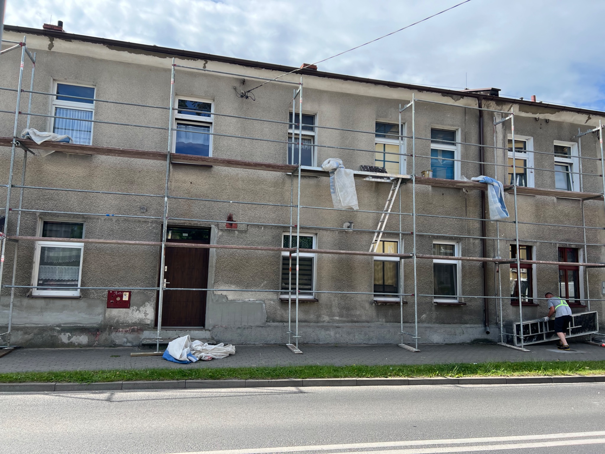 Dwukondygnacyjny budynek mieszkalny w trakcie prac elewacyjnych, otoczony rusztowaniem, z widocznymi śladami napraw i przygotowań do malowania, pracownik przenosi element rusztowania.