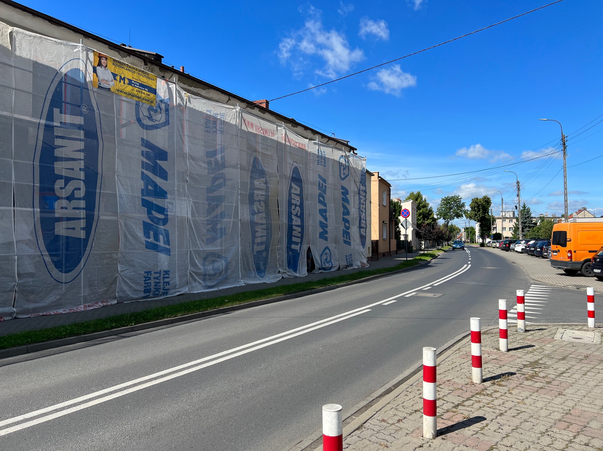Budynek w trakcie prac elewacyjnych, osłonięty siatką z nadrukiem reklamowym firm Arsanit i Mapei, widok z ulicy w słoneczny dzień.