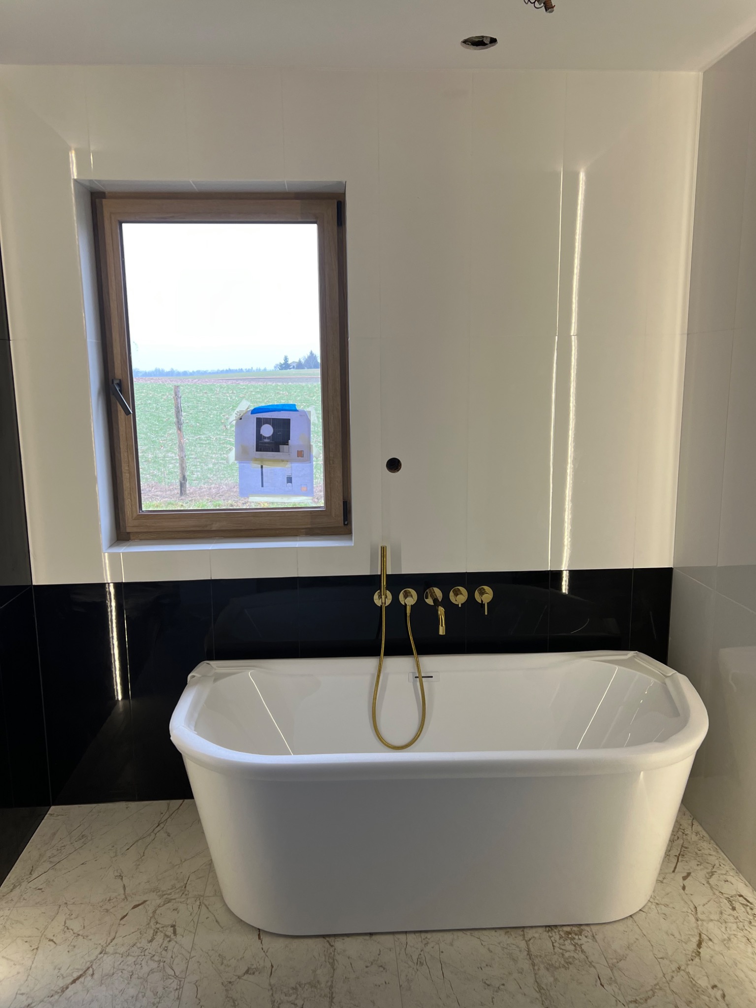 Nowoczesna łazienka w trakcie wykańczania: biała wanna wolnostojąca z złotą armaturą na tle białych płytek z czarnym pasem, widok na pole przez okno z zawieszonym rysunkiem domu.