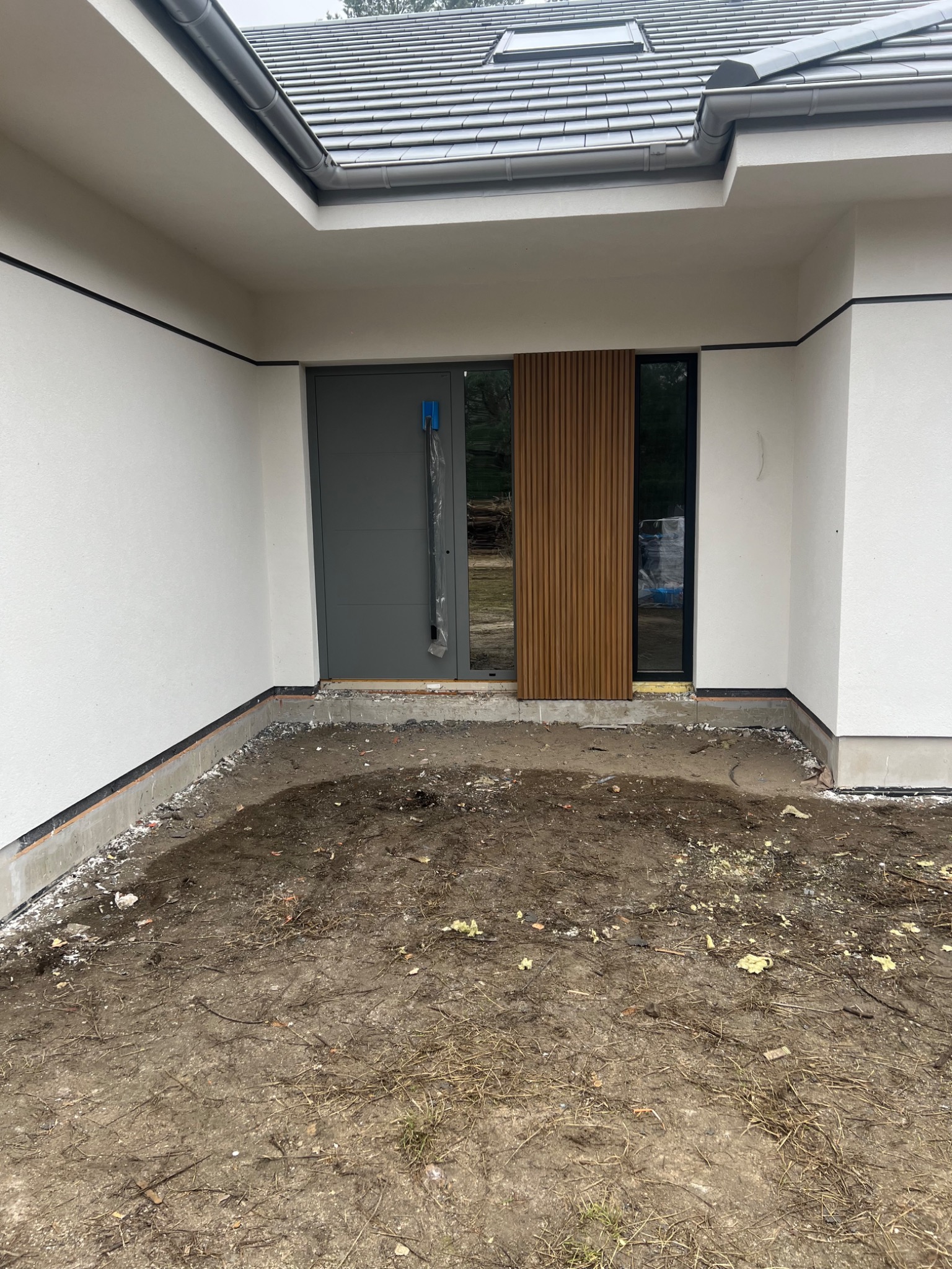 Nowoczesny dom jednorodzinny z szarą stolarką okienną i drewnianą okładziną elewacyjną przy wejściu, w trakcie prac wykończeniowych.