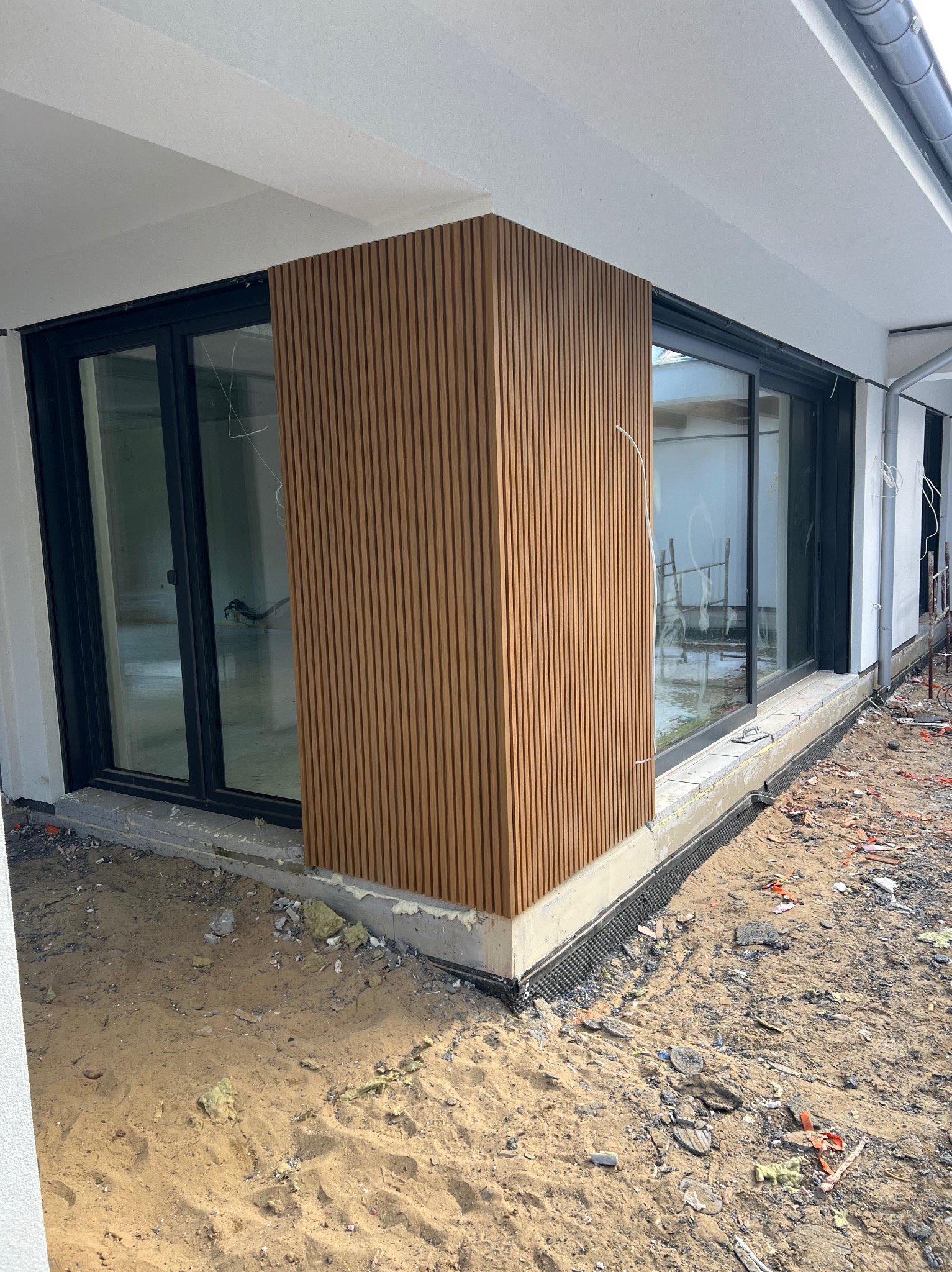 Nowoczesna elewacja budynku z drewnianymi panelami pionowymi, widok z zewnątrz, teren budowy w tle. Detal wykończenia narożnika budynku.