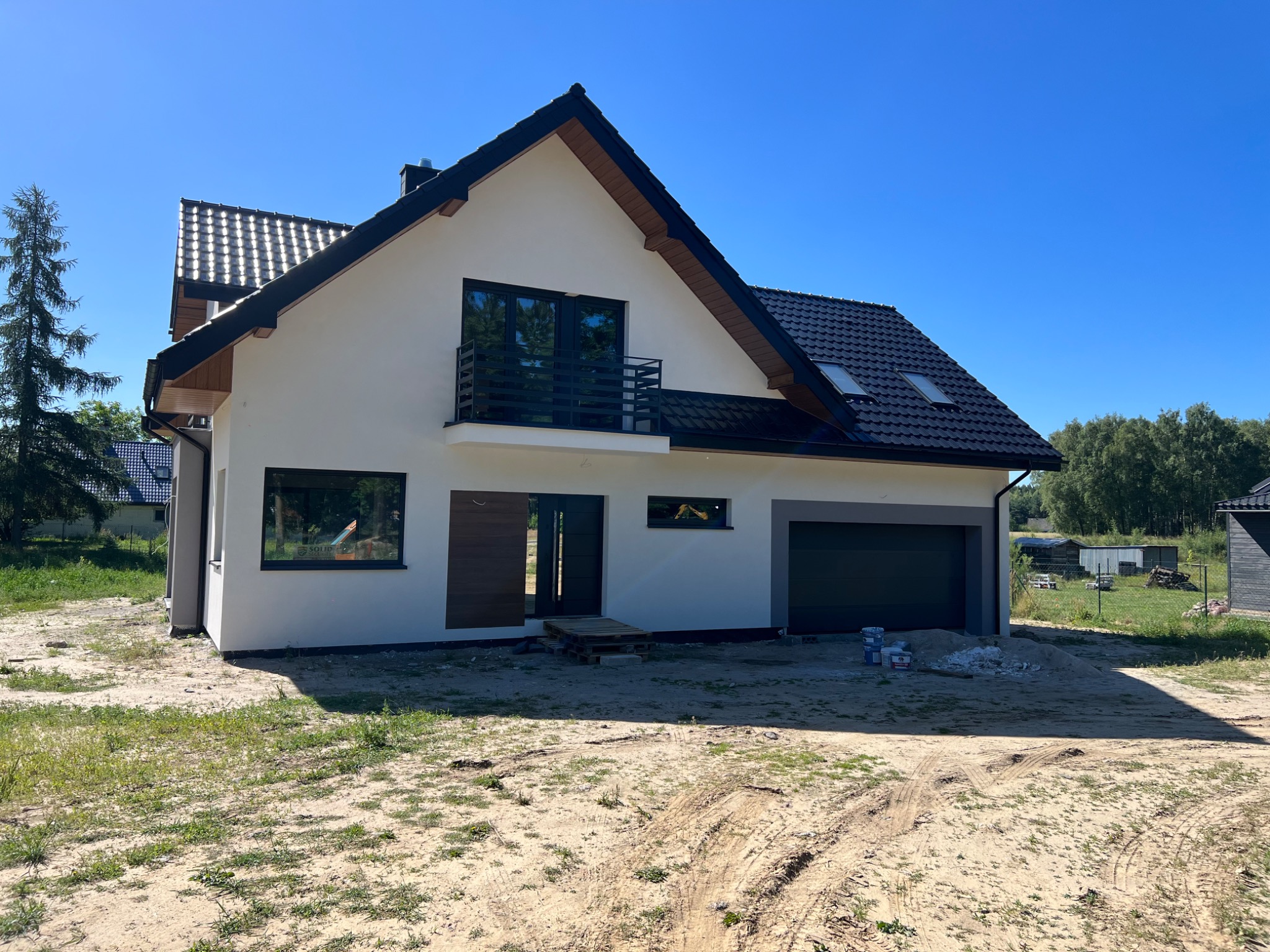 Nowoczesny dom jednorodzinny z balkonem i garażem, świeżo po malowaniu elewacji na jasny kolor, z ciemną dachówką i stolarką okienną. Widoczne puszki po farbie.