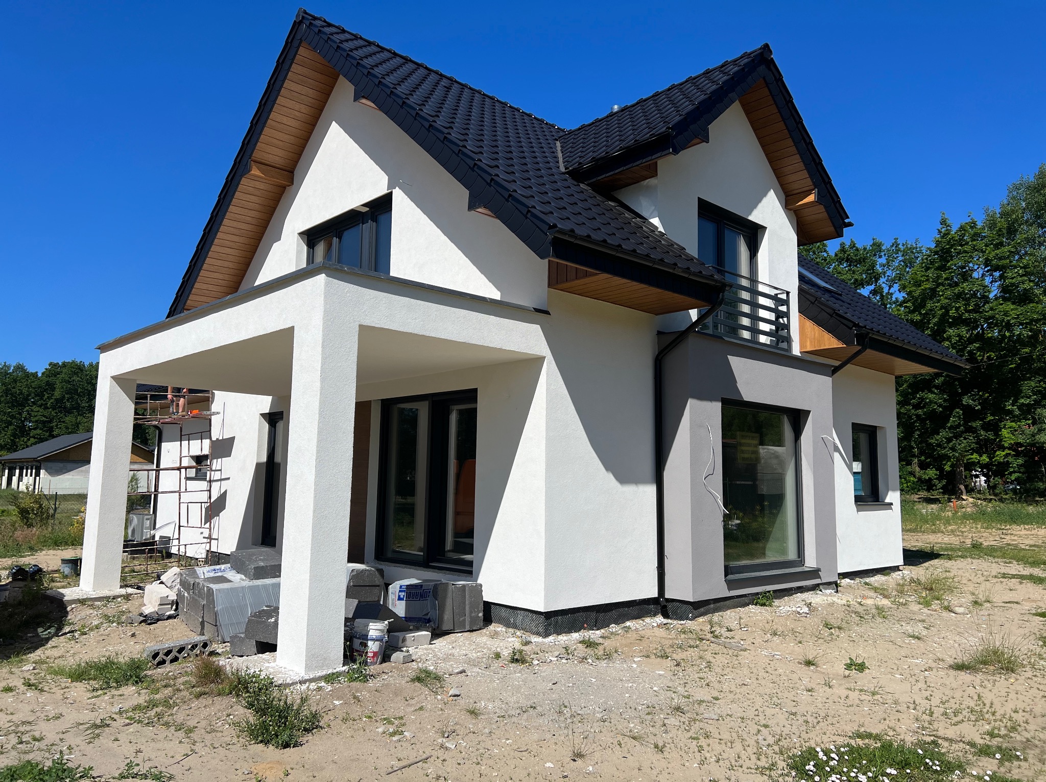 Nowoczesny dom z białą elewacją i czarnym dachem, widoczny z zewnątrz, z elementami szarości i drewnianymi akcentami. Na pierwszym planie materiały budowlane.