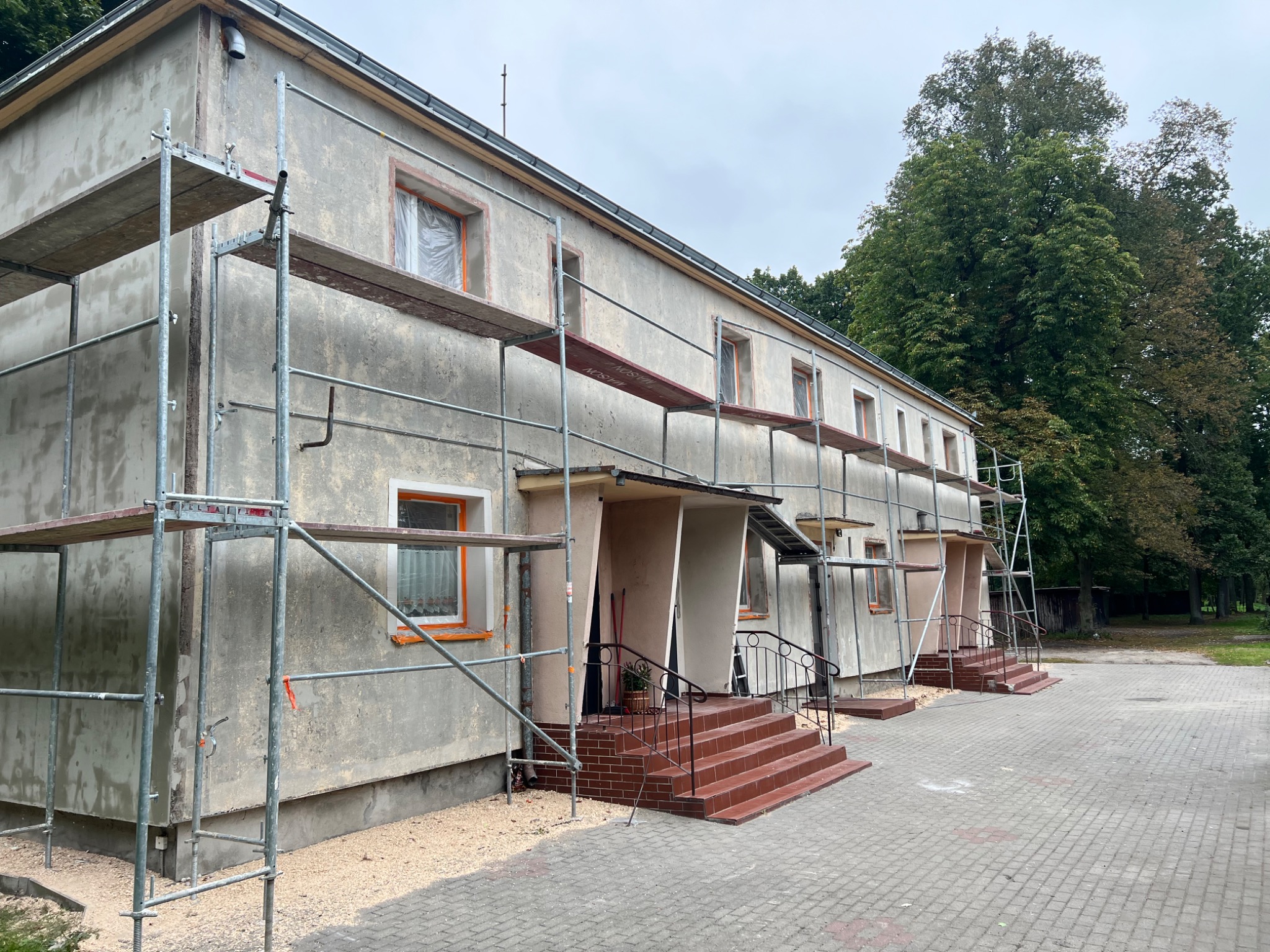 Budynek dwukondygnacyjny w trakcie prac elewacyjnych. Widoczne rusztowania, zabezpieczone okna folią i świeżo nałożony tynk. Częściowo wykończone schody wejściowe.