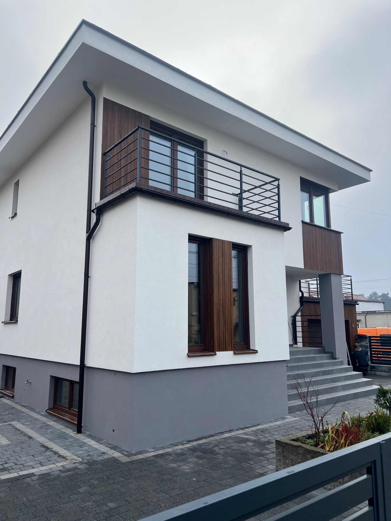 Nowoczesny dom z białą elewacją, drewnianymi akcentami i szarą podmurówką. Widoczny balkon z metalową balustradą i schody wejściowe. Minimalistyczny design.