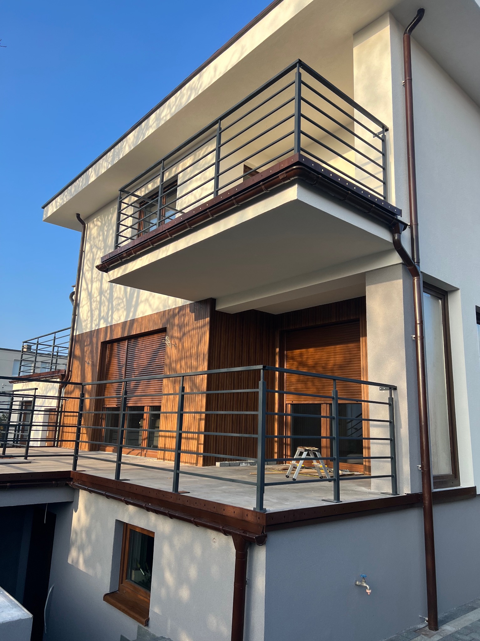 Nowoczesny dom z balkonami i drewnianą elewacją, oświetlony popołudniowym słońcem. Widoczne detale wykończenia i elementy architektoniczne.