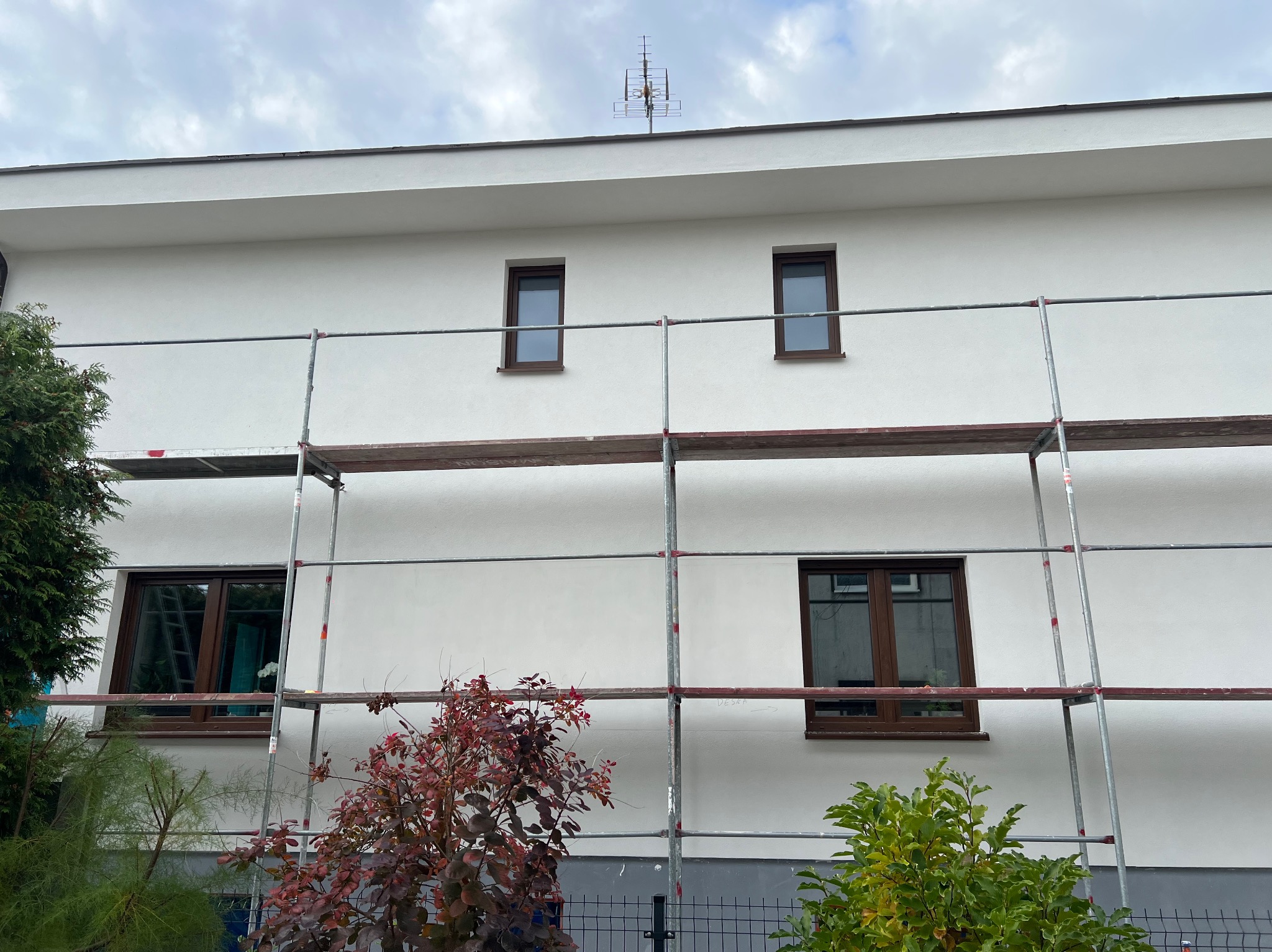Biała elewacja domu z brązowymi oknami, częściowo zasłonięta rusztowaniem. Widoczne krzewy i drzewa w tle. Antena na dachu. Pochmurne niebo.