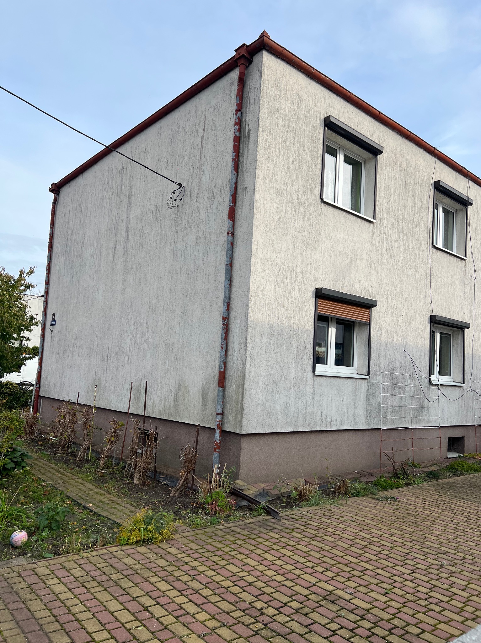Dwupiętrowy dom z szarą elewacją i brązowym cokołem, widoczne okna z roletami, rynny z śladami korozji, kostka brukowa i zaniedbana zieleń wokół budynku.