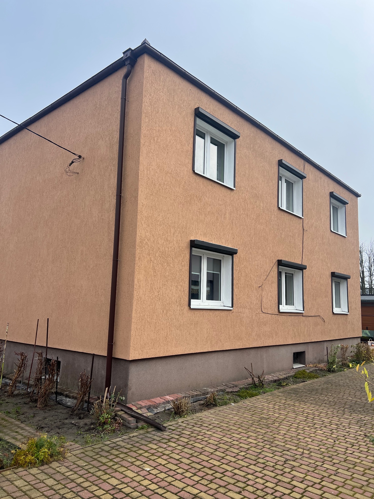 Dwukondygnacyjny dom z beżową elewacją i brązowymi rynnami, widoczny z perspektywy chodnika z kostki. Okna z roletami zewnętrznymi. Niska podmurówka.