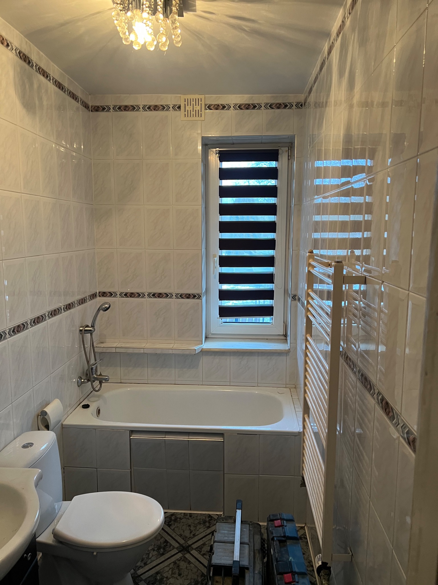 Jasna łazienka z wanną, toaletą i grzejnikiem drabinkowym. Widoczne płytki z dekoracyjnym pasem i okno z roletami dzień-noc. Częściowo widoczne narzędzia.