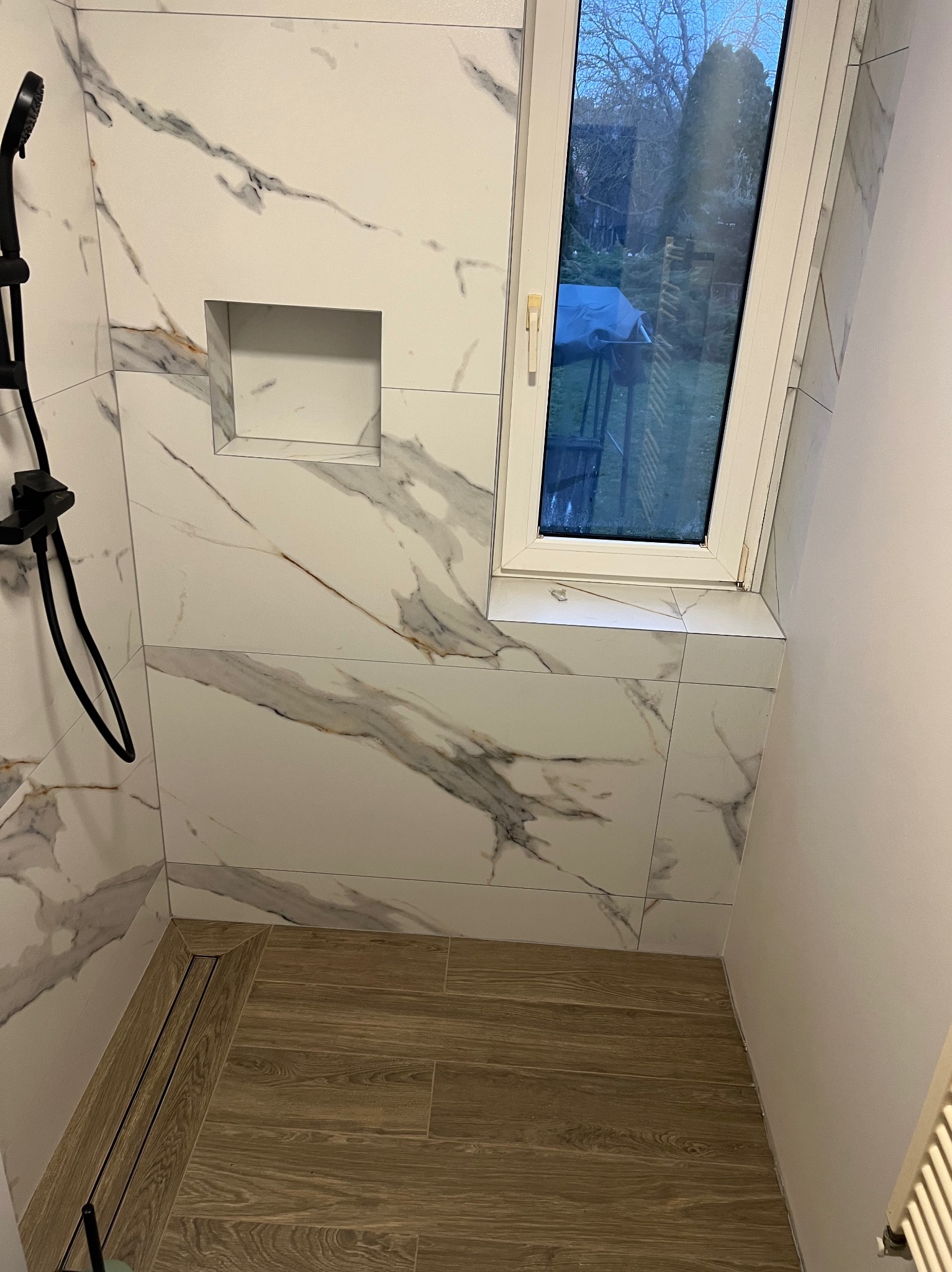 Wykończona kabina prysznicowa z marmurowymi płytkami, wnęką na kosmetyki i odpływem liniowym. Widoczne okno dachowe i drewnopodobna podłoga.