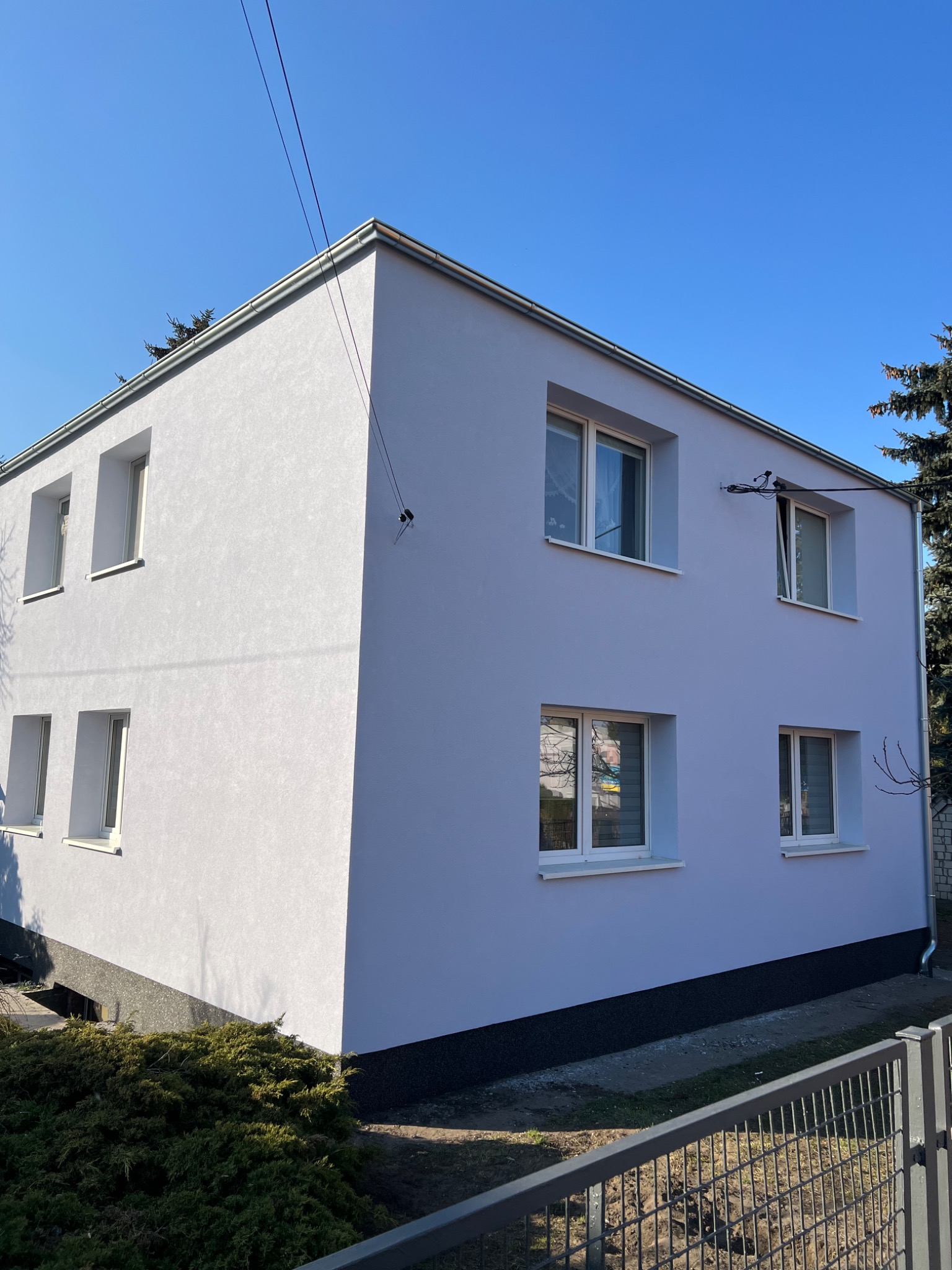 Dwukondygnacyjny dom z nową, jasnoszarą elewacją i białymi oknami, sfotografowany w słoneczny dzień z perspektywy ogrodu. Widoczne krzewy i fragment ogrodzenia.