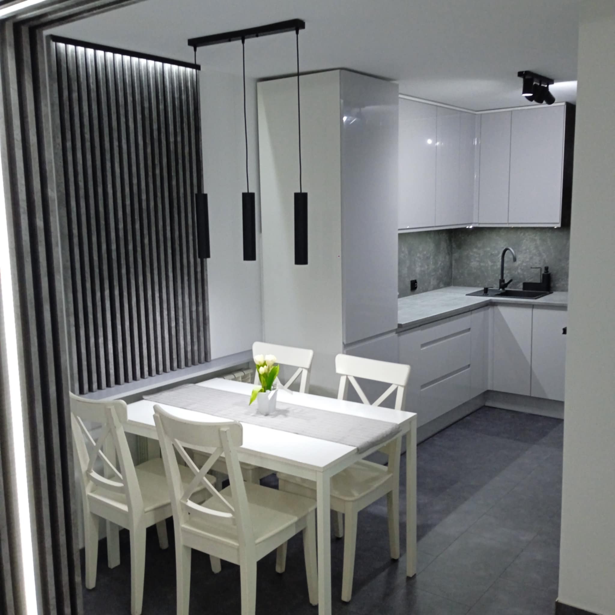 Nowoczesna aranżacja wnętrza: biała kuchnia z jadalnią, minimalistyczne lampy wiszące, dekoracyjne panele ścienne i białe krzesła przy stole z kwiatami.