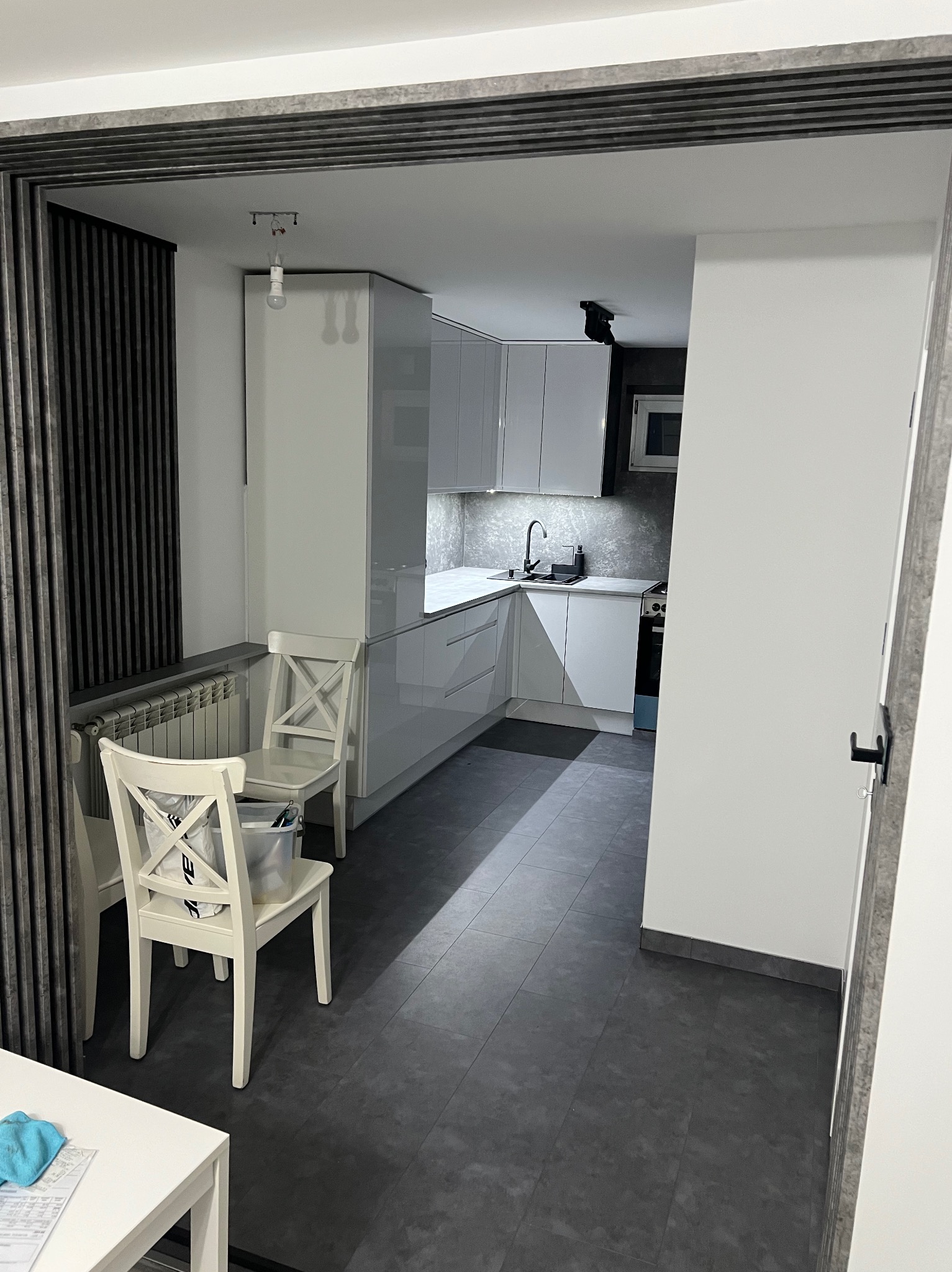 Nowoczesna, jasna kuchnia z białymi meblami i szarą podłogą, widziana z perspektywy pokoju. Uwagę zwracają białe krzesła i dekoracyjne panele ścienne.