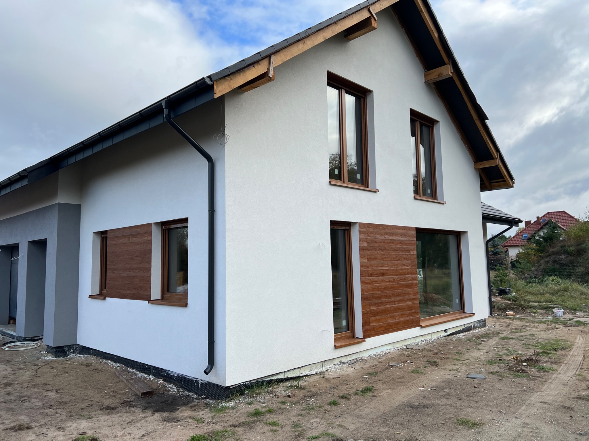 Nowoczesny dom jednorodzinny z białą elewacją i drewnianymi akcentami okiennymi oraz częściową okładziną imitującą drewno, widoczne elementy drenażu i teren w trakcie zagospodarowywania.